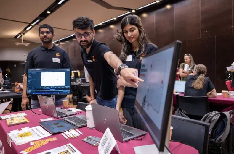 Gaurav at ASU AI Day, pointing at a monitor while explaining how LIVE builds a video, with Nishad Patne and a teammate beside him at the LEI table.